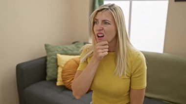 Şaşırmış ve büyülenmiş, evdeki tişörtlü genç sarışın kadın, elleri çenesinde inançsızlık - merak ve şaşkınlık ifadesi.