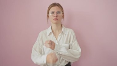 Üzgün genç sarışın kadın, şüpheci şüpheci, gözlük takan, kaşlarını çatan. Olumsuz insan, gergin ve hoşnutsuz, pembe arka planda izole edilmiş. 