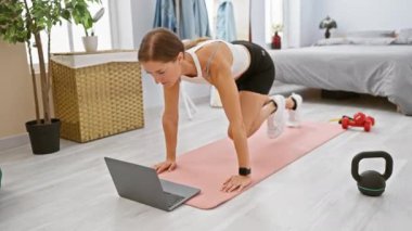 Odaklanmış genç bir kadın odasında egzersiz yapıyor, dizüstü bilgisayarındaki egzersiz videosunun ardından, spor ekipmanlarıyla çevrili..