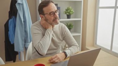 Orta yaşlı, gözlüklü ve sakallı bir adam. Modern bir evde dizüstü bilgisayarla çalışırken boynu ağrıyor..