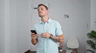 Sakallı ve dövmeli genç İspanyol bir adam modern ev ortamında akıllı telefon tutuyor. Bekleme ya da beklenti hissi veriyor..