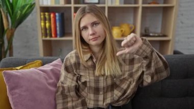 Kızgın genç sarışın kadın evde, olumsuz jestler yapıyor. Kanepede otururken mutsuz bir ifade..
