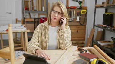 Güvenlik gözlüklü kendine güvenen bir kadın marangozluk atölyesinde telefonla konuşuyor. Etrafı aletlerle ve odunlarla çevrili..