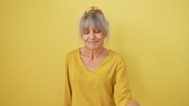 Midilli kuyruklu neşeli orta yaşlı kadın, merhaba diyor, neşeli, dostane bir hoş geldin jesti. İzole edilmiş sarı arka planda mutlu bir gülümseme.