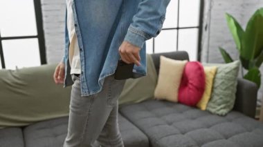 Kot ceketli genç bir adam renkli yastıklı rahat bir oturma odasında akıllı telefon kullanıyor..