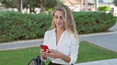 Büyüleyici bir portre, genç sarışın kadın ayağa kalkıyor, el sallıyor ve merhaba derken yeşil parkta akıllı telefon kullanıyor.