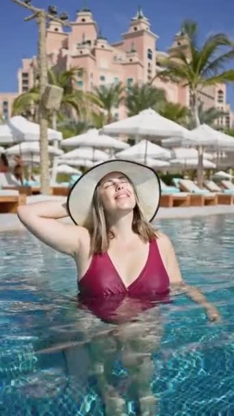 Maroon 수영복과 Sunhat에서 여자는 배경에 손바닥과 카바나와 호화로운 리조트 수영장에서 — 비디오