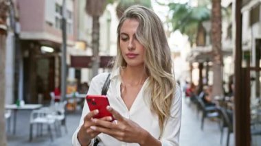 Çekici sarışın bir kadın, tamamen dalgın, şehrin güneşli ufuk çizgisinin altındaki sıcak bir kahve dükkanının terasında telefonuyla mesajlaşıyor.