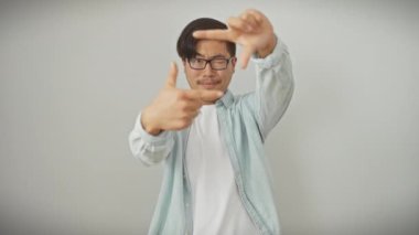 Gözlüklü mutlu yüzlü genç Çinli bir adam, bir vizyon tasarlıyor ve izole edilmiş beyaz arka planda el hareketleriyle bir fotoğraf çerçeveliyor.