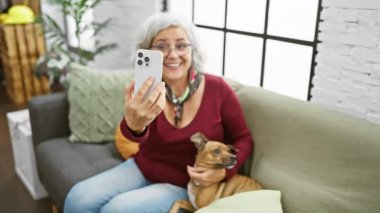 Gözlüklü, gri saçlı, gülümseyen olgun bir kadın köpeğiyle rahat bir oturma odasında selfie çeker..