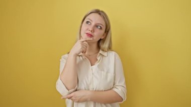 Sarışın genç bir kadın, yüzünde ciddi bir ifade, çenesinde el, düşünceli bir şekilde duruyor, kafa karıştırıcı bir fikir düşünüyor, sarı bir arka planda izole edilmiş.