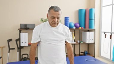 Tam donanımlı bir fizyoterapi kliniğinde, rehabilitasyon sürecini göz önünde bulundurarak düşünceli bir şekilde ayakta duran olgun bir İspanyol..