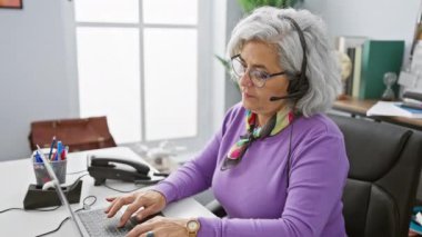 Modern ofiste dizüstü bilgisayarda çalışan, güler yüzlü, gri saçlı bir kadın profesyonelliği ve arkadaşlığı yansıtıyor..