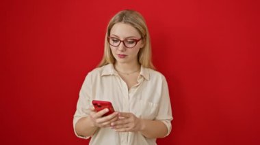 Neşeli genç sarışın kadın, akıllı telefonuyla mesajlaşırken izole edilmiş kırmızı arka plan üzerinde 
