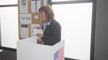 Amerikan seçimlerinde oy kullanan orta yaşlı bir kadın bizimle birlikte oy kullanma merkezinde bayrak taşıyor.