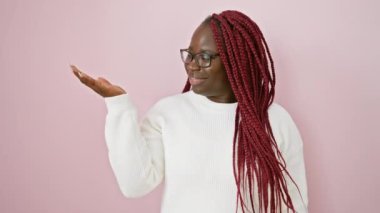 Gözlüklü ve beyaz kazaklı gülümseyen Afrikalı bir kadın pembe bir arka plana uzanmış bir elle poz veriyor..