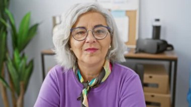 Gözlüklü ve gri saçlı gülümseyen kadın ofis ortamında poz veriyor, profesyonelliği ve deneyimi simgeliyor..