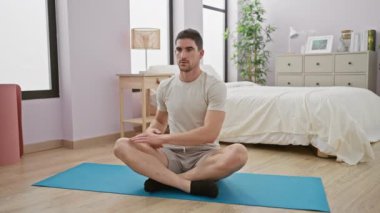 Parlak, çağdaş bir yatak odasında yoga minderinin üzerinde bağdaş kurmuş, huzurlu bir atmosferi ifade eden günlük kıyafetli bir adam..