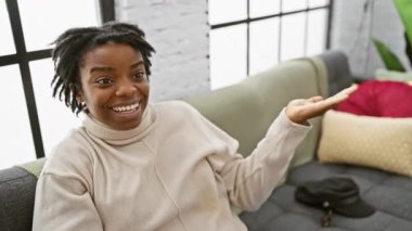 Genç siyah kadın kanepeye oturmuş neşeli bir şekilde gülümsüyor ve avuç içi kameraya bakıyor. evde.