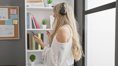Genç sarışın bir kadın, modern ofis ortamında elinde akıllı bir telefonla kulaklıkla müzik dinlemekten hoşlanıyor..