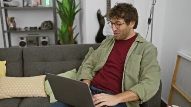 Cheerful young man with glasses and a beard smiling while using a laptop and holding a heart-shaped box in a cozy living room.