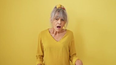 Şok olmuş orta yaşlı kadın, at kuyruklu gri saçlı, yüzünde hayret verici bir ifade sergiliyor, izole edilmiş sarı arka planda korku ve şaşkınlık ifadesi sergiliyor..