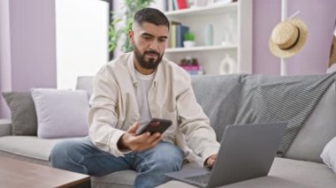 Sakallı bir adam akıllı bir telefonla konuşuyor ve modern oturma odasında rahat bir şekilde otururken dizüstü bilgisayar kullanıyor. Sıradan bir yaşam tarzı ve uzak bir çalışma ortamı sergiliyor..