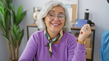 Neşeli, gri saçlı, orta yaşlı kadın, elinde iyimser bir gülümseme ve açık ağızlı bir ifadeyle, güvenle baş parmağını ofisin içine uzatıyor! pozitif finans fikri!