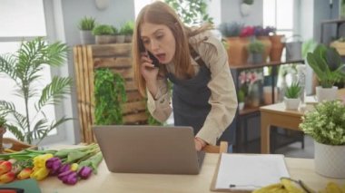 Odaklanmış genç, kızıl saçlı bir kadın botanik dolu bir iç mekanda bir telefon görüşmesi ve dizüstü bilgisayarla birden fazla iş yapıyor..