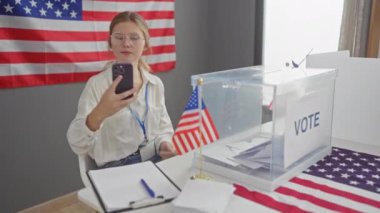 Genç bir kadın bir Amerikan oy kullanma merkezinde Amerikan bayrağı ve oy sandığıyla selfie çekiyor..
