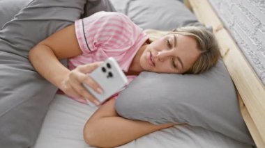 Genç bir kadın rahat bir yatak odasında akıllı bir telefonla yatakta dinleniyor..