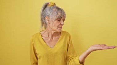 Neşeli orta yaşlı, at kuyruklu, gri saçlı kadın mutlu bir şekilde avucuyla bir şey gösteriyor, güneşli, izole edilmiş sarı bir arka planda kameraya bakarken gülümsüyor.