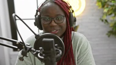 Kırmızı örgülü neşeli bir Afrikalı kadın profesyonel bir radyo stüdyosunda podcast yapıyor..