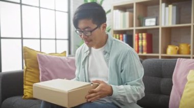 Gülümseyen Asyalı bir adam, modern bir oturma odasında otururken bir paketi açıyor, neşe ve merakını dile getiriyor..