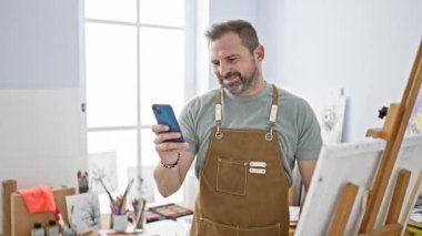 Beyaz saçlı, yakışıklı, orta yaşlı bir adam. Parlak bir sanat stüdyosunda akıllı telefon kullanıyor.