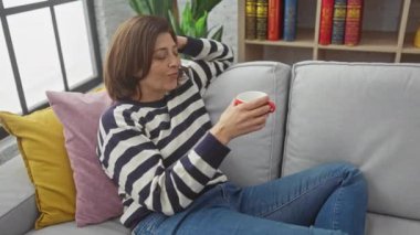 Rahat orta yaşlı bir kadın şık bir oturma odasında rahat bir kanepede kahve içiyor..