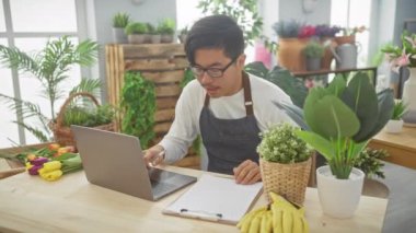 Kapalı bir çiçekçide dizüstü bilgisayarda çalışan Asyalı genç bir adam çeşitli bitki ve çiçeklerle çevrili..