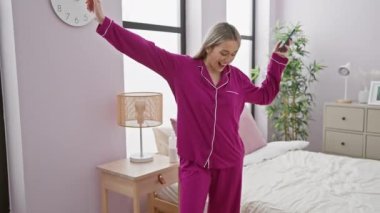 Canlı pembe pijamalı neşeli genç bir kadın modern bir yatak odası ortamında elinde akıllı bir telefonla dans ediyor. Mutluluk ve konfor ifade ediyor..