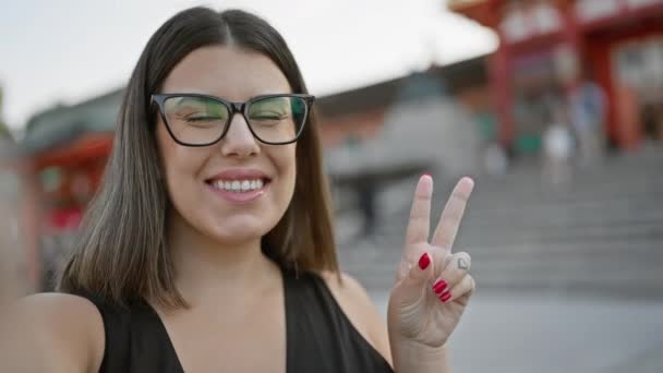楽しくカメラにメガネの波で自信を持って幸せなヒスパニック女性 京都の長見稲荷大社からのカジュアルなビデオ通話 — ストック動画