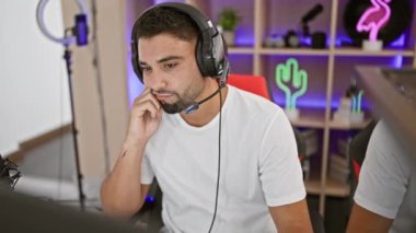 Kulaklığı olan odaklanmış bir adam renkli bir oyun odasında oturup bilgisayar ekranına bakar, bağlılığı ve konsantrasyonu yansıtır..