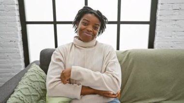 Kendinden emin genç siyah bir kadın. Rastalı saçları parıldayan yüzünü çerçeveliyor. Oturma odasındaki rahat bir kanepeye kurulmuş. Elleri yanda, bulaşıcı gülümsemesi yüzünde parlıyor..