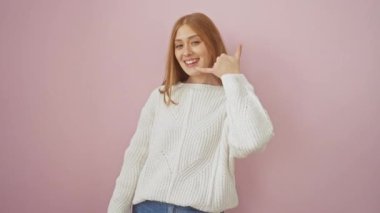 Neşeli, genç, kızıl saçlı bir kadın parmaklarıyla bir telefon konuşması yapıyor, kazak giyiyor, pembe izole bir zemin üzerinde, pozitif iletişim titreşimleri yayıyor.