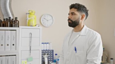 Beyaz laboratuvar önlüklü, sakallı, genç, Latin asıllı, düşünceli bir erkek bilim adamı..