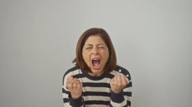 Kızgın İspanyol kadın, çizgili kazak giyiyor, agresif bir ifadeyle çılgınca bağırıyor ve soyutlanmış beyaz arka plan yüzünden hayal kırıklığı içinde kollarını kaldırıyor.