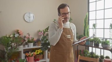 Önlüklü olgun bir adam çiçeklerle dolu bir çiçekçide elinde bir pano tutarken telefonla konuşuyor..