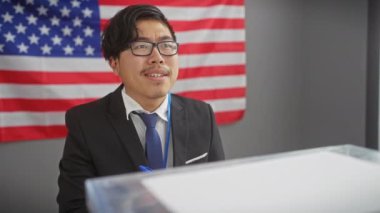 Takım elbiseli genç bir Asyalı podyumda konuşuyor. Kapalı alanda Amerikan bayrağı var..