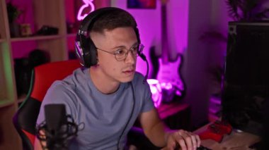 Kulaklık ve gözlük takan odaklanmış bir adam geceleri renkli, neon ışıklı bir oyun odasında bilgisayar kullanıyor..