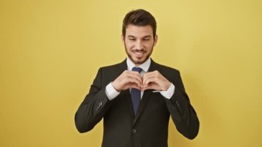 Takım elbiseli neşeli İspanyol bir adam, elleri ile bir kalp sembolü yapar, sarı izole bir arka plana karşı durur, romantik bir jestle aşkla gülümser.