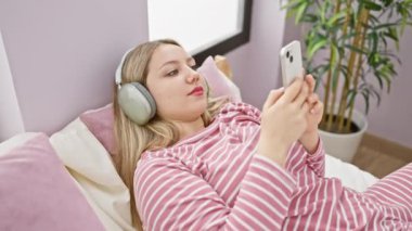 Kulaklık takan rahat genç bir kadın rahat, bitki süslemeli bir yatak odasında akıllı telefonunu karıştırıyor.