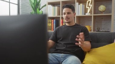 Sakallı, siyah tişörtlü genç bir İspanyol koltukta dinleniyor, oturma odasında televizyon seyrediyor..